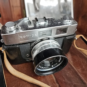 Puede incluir: Una cámara Konica vintage con una lente negra y un cuerpo plateado. La cámara tiene una correa de cuero y está en una funda de cuero negra. La lente tiene marcas de medición en pies. La palabra "Konica" está impresa en el cuerpo de la cámara.