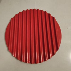 May include: A red, round, silicone trivet with a corrugated surface. The trivet is designed to protect surfaces from heat.