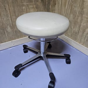 May include: A white, round, adjustable stool with a chrome base and black wheels. The stool has a padded seat and is designed for comfort and mobility. The base has five legs for stability.