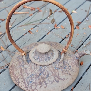 May include: A round, flat-bottomed ceramic teapot with a textured, sandy-beige surface featuring a dark brown lace-like pattern and small blue accents. It includes a matching circular lid and a tall, arched handle made of natural brown bamboo.