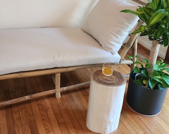 May include: A white wooden stool with a round top and a natural wood base. A small glass with a yellow liquid sits on the stool. The stool is in front of a white couch with a light gray cushion.