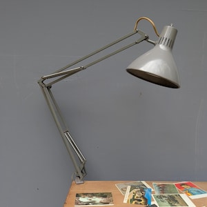 May include: A grey articulated desk lamp with a conical shade and a clamp base. The lamp features a long, adjustable metal arm with visible hinges and a light-colored cord loop at the top. The shade has a series of vertical vents near the top.