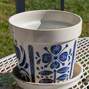 May include: A ceramic flower pot with a matching saucer. The pot is off-white with a blue floral pattern. The pot is sitting on a white metal table.