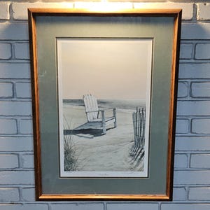 May include: Framed print of a beach scene with a white Adirondack chair on the sand. The artwork features a wooden frame, a green mat, and a serene coastal landscape with a fence and ocean in the background.