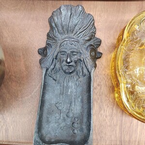 May include: A vintage metal ashtray featuring a Native American chief in a headdress. The ashtray is rectangular with a detailed relief of the chief's face and headdress. The color is a dark, aged metal, and it sits on a wooden surface.