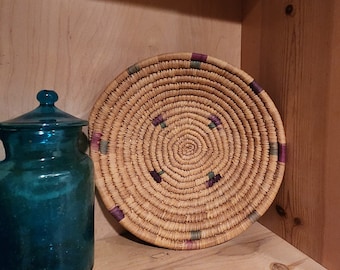 May include: A round woven basket with a natural straw color and purple and green accents. The basket is sitting on a wooden shelf.