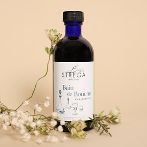 May include: A blue glass bottle with a black cap, labeled "STREGA della Veda Bain de Bouche aux plantes HERBORISEE". The bottle is sitting on a light beige surface with white flowers.