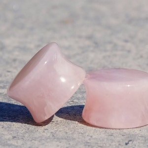 Puede incluir: Dos tapones para los oídos de cuarzo rosa. Uno está en ángulo, mostrando una forma ensanchada y redondeada. El otro está en posición vertical, mostrando una forma cilíndrica con un borde ensanchado. Ambos son de color rosa pálido.