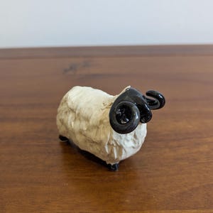 May include: A small figurine of a ram with a textured, off-white body and glossy, black curled horns. The ram stands on four small, dark-coloured hooves, showcasing a detailed, woolly coat texture and a distinct, curved horn structure.