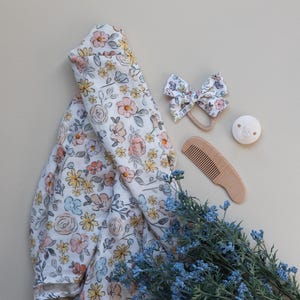 May include: A floral print baby swaddle blanket, a wooden comb, a wooden teether, and a floral print hair bow. The items are arranged on a light beige surface with a sprig of blue flowers.