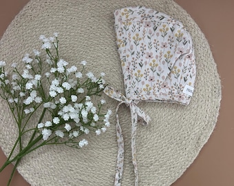 Könnte beinhalten: Ein weißer Häubchen mit einem Blumenmuster in Rosa-, Gelb- und Grüntönen. Das Häubchen hat einen Bindegurt an der Rückseite und das Wort "Peace" ist auf der Seite aufgedruckt.
