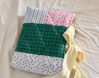 May include: A quilted fabric bag with a patchwork design. The bag features sections of green, pink, and white patterned fabric. A yellow fabric tie is attached to the side. The bag is likely used for carrying items.