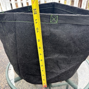 Puede incluir: Bolsa de cultivo de tela negra con costuras verdes y una correa negra. Una cinta métrica amarilla está al lado de la bolsa, midiendo 28 cm.