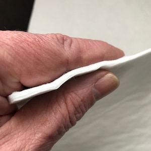 Puede incluir: Primer plano de un tejido blanco, suave y flexible. El tejido se sostiene en una mano, mostrando su textura y grosor.