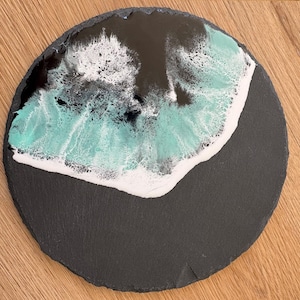 Könnte beinhalten: Runder, dunkelgrauer Schiefer-Servierteller mit einem abstrakten Design. Das Design zeigt eine Mischung aus türkis, weißem und schwarzem Harz, das an Meereswellen erinnert. Der Teller hat einen Durchmesser von etwa 25 cm.
