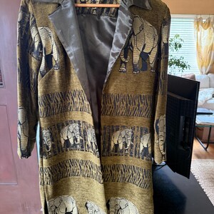 May include: A long, olive green coat with a repeating elephant pattern in gold and black. The coat has a black satin collar and lining. The coat is hanging on a wooden hanger.
