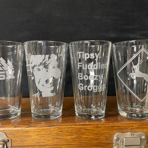 May include: Four clear glass pint glasses with different designs etched into them. The glasses are on a wooden surface. The designs include a band logo, a portrait, a list of words, and a deer silhouette.