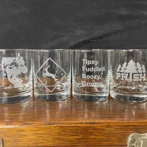 May include: Four clear glass tumblers with different etched designs. The first tumbler has a silhouette of a person's head. The second tumbler has a deer in a road sign. The third tumbler has the words "Tipsy, Fuddled, Boozy, Groovy". The fourth tumbler has the words "Phish" and a pine tree design.