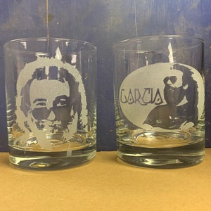 May include: Two clear glass tumblers with etched designs. One tumbler features a portrait of Jerry Garcia, the other has the word "Garcia" etched in a circle.