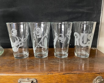 May include: Four clear glass pint glasses with etched designs. The glasses feature images of musicians and the word "Rush".