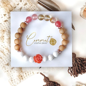 Könnte beinhalten: Ein handgefertigtes Armband mit einer Mischung aus natürlichen Elementen. Es enthält hellbraune Holzperlen, weiße poröse Perlen und farbige Edelsteinperlen. Eine korallenfarbene Rose sorgt für einen dekorativen Akzent. Die Karte trägt den Text "Cennet creative pieces".