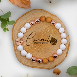 Könnte beinhalten: Ein weißes und roségoldenes Perlenarmband mit Holzperlen und einem Lebensbaum-Anhänger. Das Armband liegt auf einer Holzoberfläche mit weißem Hintergrund.