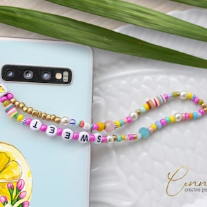 Puede incluir: Un abalorio de teléfono de cuentas coloridas con la palabra "SWEET" escrita en letras blancas sobre cuentas rosas. El abalorio está unido a una funda de teléfono azul claro con un diseño de limón.