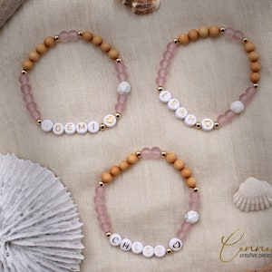 May include: Three personalised bracelets with wooden and pink glass beads. The bracelets spell out the names "Noemi", "Leah", and "Chloe" in gold lettering.