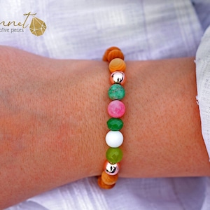 Könnte beinhalten: Ein Armband mit bunten Perlen aus Holz und Edelsteinen. Das Armband enthält orange, grüne, rosa und weiße Perlen, getrennt durch silberfarbene Metallperlen. Das Armband wird am Handgelenk getragen.