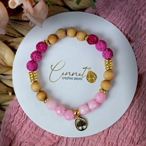 Könnte beinhalten: Ein rosa-braunes Perlenarmband mit einem goldenen Baum-Anhänger. Das Armband besteht aus Lava-Perlen, Holzperlen und Rosenquarz-Perlen. Der Anhänger ist ein goldener Baum mit einer kleinen, runden, goldenen Perle an der Spitze.