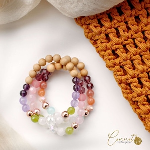 Könnte beinhalten: Ein mehrfarbiges Armband mit Holzperlen, roségoldenen Perlen und verschiedenen Edelsteinperlen in den Farben Rosa, Lila, Grün und Orange. Das Armband liegt auf einem weißen Hintergrund.