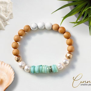 Könnte beinhalten: Ein Armband aus Holzperlen, weißen und türkisfarbenen Steinen und goldenen Akzenten. Das Armband liegt auf einem weißen Hintergrund mit Muscheln.