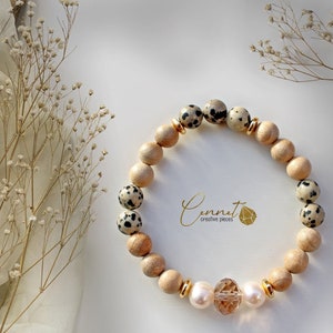 Puede incluir: Un brazalete hecho con cuentas de madera marrón y cuentas de jaspe dalmata blanco y negro. El brazalete tiene un acento dorado y una gran cuenta de cristal en el centro.
