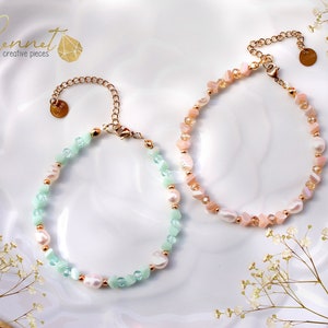 May include: Two bracelets with gold-coloured chains and clasps. One bracelet features pale green and white beads, the other has pink and white beads. Both bracelets have a small gold charm.