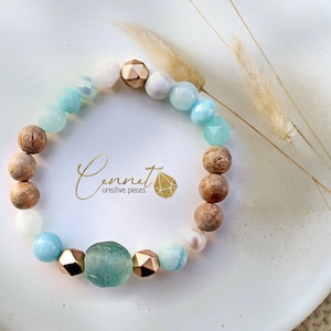 Könnte beinhalten: Ein hellblaues, weißes und braunes Perlenarmband mit goldenen Akzenten. Das Armband besteht aus verschiedenen Perlen, darunter Holz, Achat und Glas. Das Armband liegt auf einer weißen Oberfläche mit getrocknetem Gras.