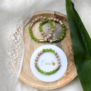 Può includere: Due braccialetti di legno e perline verdi con dettagli dorati su un vassoio di legno. I braccialetti sono impilati uno sopra l'altro. Il vassoio è su una tovaglia di pizzo bianco. Il testo "Connect Creative Pieces" è su un biglietto bianco al centro del vassoio.