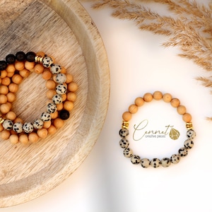 Könnte beinhalten: Zwei Armbänder aus natürlichen Holzperlen und Dalmatiner-Jaspisperlen. Die Armbänder sind auf einem Holzteller gestapelt. Das kleinere Armband hat goldene Akzente. Das größere Armband hat schwarze Lavasteine und goldene Akzente. Der Text "Connect creative pieces" ist im Bild sichtbar.