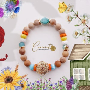 Könnte beinhalten: Ein farbenfrohes Holz-Armband mit einem goldenen Filigranverschluss. Das Armband besteht aus orangefarbenen, gelben, weißen und blauen Perlen sowie einem türkisfarbenen Stein. Das Armband ist von Blumen und einem kleinen Haus umgeben.