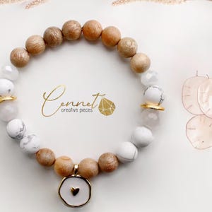 Può includere: Un braccialetto realizzato con legno e perline bianche con un ciondolo a forma di cuore dorato. Il braccialetto ha una chiusura dorata ed è accentato da distanziatori dorati. Il braccialetto è su uno sfondo bianco.