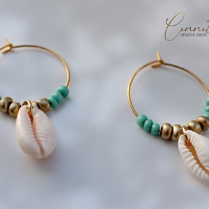 Puede incluir: Aretes de aro dorados con un pequeño dije de concha blanca y cuentas turquesas.