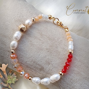 Puede incluir: Un brazalete dorado con cuentas blancas, melocotón y rojas. El brazalete tiene un cierre dorado y una cuenta geométrica dorada.