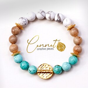May include: A turquoise, white, and brown beaded bracelet with gold accents. The bracelet features a large, hammered gold charm in the center.