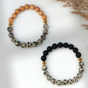 May include: Two bracelets, one with brown wood beads and the other with black lava beads, both with white and black dalmatian jasper beads and gold accents.