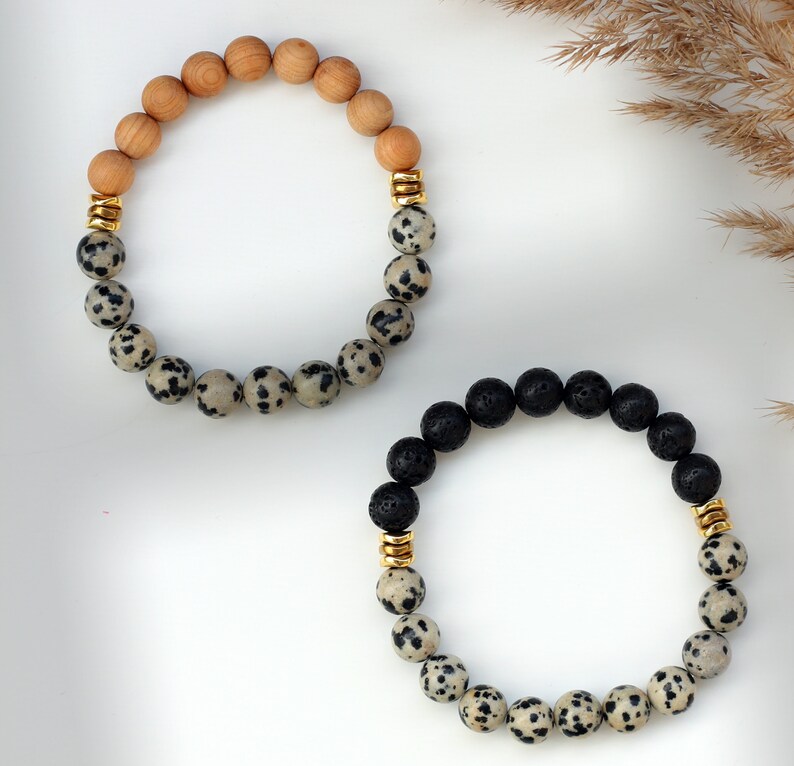 May include: Two bracelets, one with brown wood beads and the other with black lava beads, both with white and black dalmatian jasper beads and gold accents.