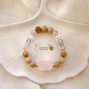 Könnte beinhalten: Ein Armband mit einem großen, rechteckigen Rosenquarzstein, Holzperlen und irisierenden und rosa Kristallperlen. Das Armband wird auf einer weißen, blattförmigen Schale präsentiert. Der Text "Cennet creative pieces" ist sichtbar.