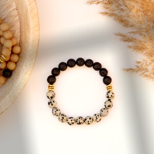 May include: A black and white speckled stone bracelet with gold accents. The bracelet is made of lava rock and dalmatian jasper beads.