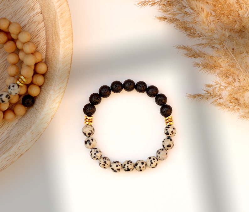 May include: A black and white speckled stone bracelet with gold accents. The bracelet is made of lava rock and dalmatian jasper beads.