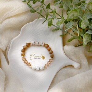 Könnte beinhalten: Ein Armband aus braunem Holz und weißen Perlen mit goldenen Akzenten und klaren Kristallperlen. Das Armband liegt auf einer weißen Keramikplatte mit dem Text "Cennat Creative Pieces".