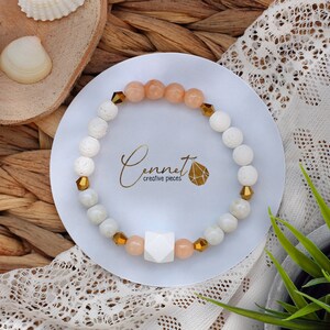 Könnte beinhalten: Weißes und pfirsichfarbenes Perlenarmband mit goldenen Akzenten und einem weißen geometrischen Anhänger. Das Armband liegt auf einem weißen Teller mit dem Text "Cennet creative pieces" darauf.