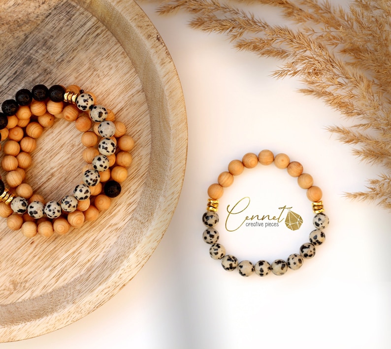 May include: Two bracelets made of natural wood beads and dalmatian jasper beads. The bracelets are stacked on a wooden plate. The smaller bracelet has gold accents. The larger bracelet has black lava beads and gold accents. The text "Connect creative pieces" is visible in the image.